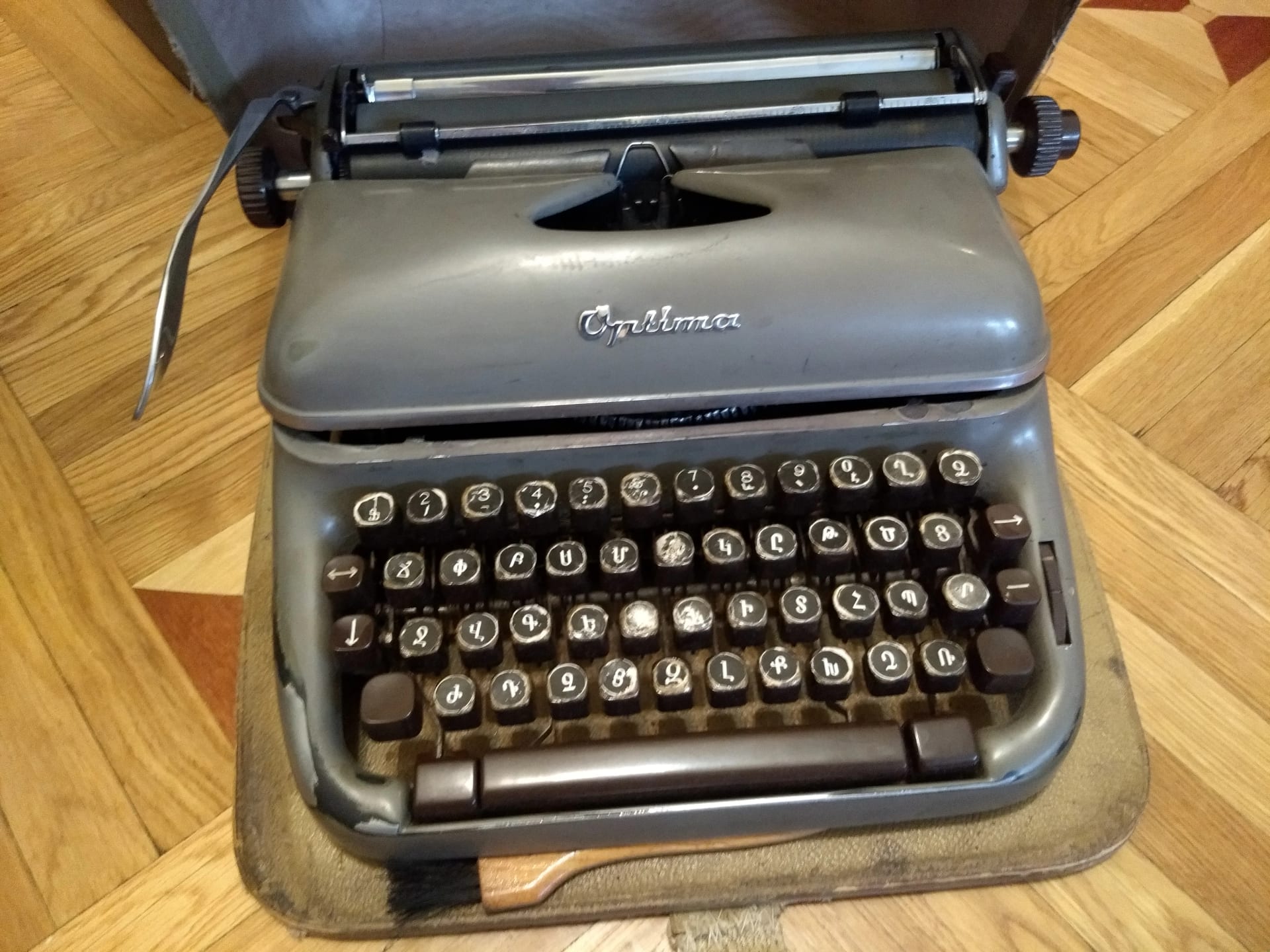 Máquina de escrever mecânica cinza, com teclado em armênio, dentro de sua maleta de couro sobre piso de madeira.